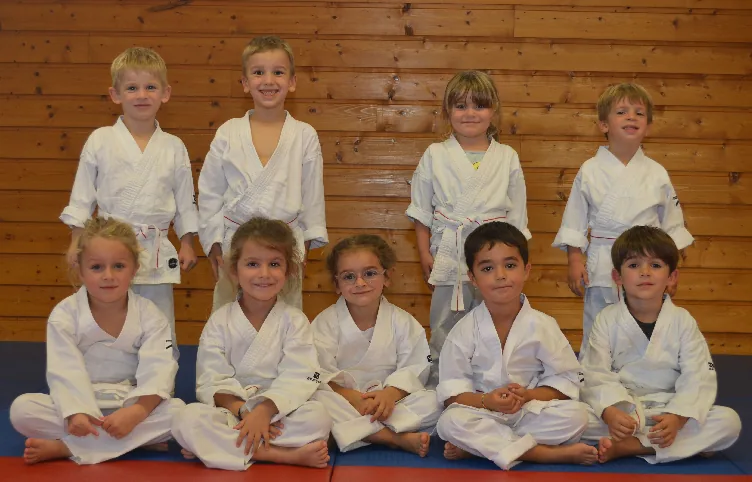 Neuf enfants du cours de Baby Karaté de Montbonnot en kimono blanc, assis et debout sur le tatami devant le mur de bois du dojo